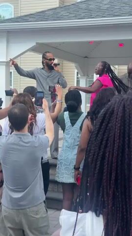 The video shows a group of people gathered outdoors, seemingly at a party or event. Three individuals are standing on a raised platform, possibly a deck or stage. The person in the center, a woman wearing a bright pink top and glasses, is holding her arms up and appears to be speaking or singing into a microphone that is not visible. To her left, a man in a gray patterned shirt and sunglasses is also holding a microphone and gesturing with his left hand, suggesting he is performing. Another man stands to the right of the woman. A crowd of people is in front of the platform, many of them holding up their phones, likely to record the performance. The environment appears to be a backyard, with a house visible in the background. The lighting suggests it is daytime.