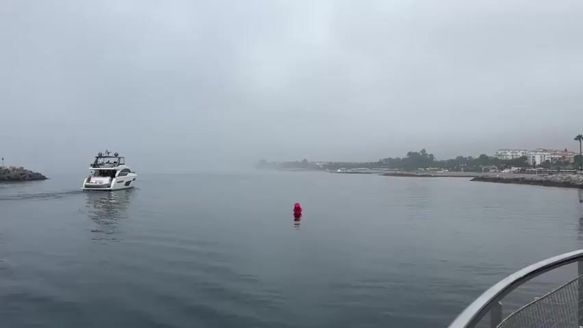 The video opens on a hazy day with a yacht sailing away from the camera into the fog. A red buoy is visible in the water to the right of the yacht. The camera then pans to the right, revealing a coastline with buildings, palm trees, and a rocky shoreline. A tall, stone tower with a red beacon at the top stands prominently on the right side of the frame, with a small, ornate white building next to it. People are visible walking along the waterfront. The water is calm, reflecting the overcast sky.