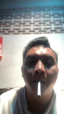 A person is holding a cigarette in their mouth. A lighter is being used to light the cigarette. The person is wearing a white shirt. The background is a wall with a patterned texture.