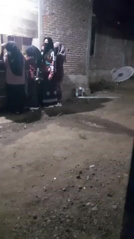 A group of about five people, likely girls or young women, are standing outside a brick building at night. The girls are wearing long dresses or skirts and headscarves, which suggest they might be Muslim. They seem to be gathered around a brightly lit area, possibly a window or a small store. The ground is dirt and there is a satellite dish visible on the side of the building. The overall impression is of a simple, perhaps rural, setting.