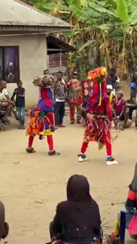 The video shows two dancers in the middle of a performance in what appears to be a village setting. They are wearing elaborate, colorful costumes, including headwear, and perform acrobatic dance moves.  The audience, consisting of men, women, and children, are sitting around the edge of the performance area, observing the dancers.  The setting is a sandy courtyard with buildings and tropical vegetation in the background. The video is shot from a medium distance, providing a clear view of the dancers and the surrounding crowd.