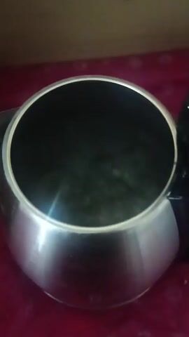 The video shows the top of a stainless steel kettle filled with water. The water is boiling and steam is rising from the surface. The kettle is sitting on a red surface, possibly a tablecloth. The background is dark and out of focus. There is no one visible in the video.