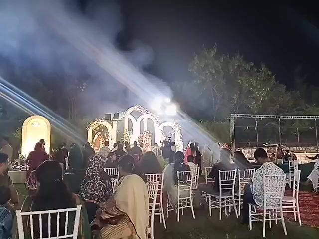 This is a video taken at night, likely outdoors. The scene appears to be a wedding ceremony or reception. There is a decorated stage with lights and smoke machines creating dramatic beams of light. A crowd of people are seated in chairs, facing the stage. Some people are standing and mingling. The ground is covered in grass, and there are trees in the background. The overall atmosphere is festive and celebratory.