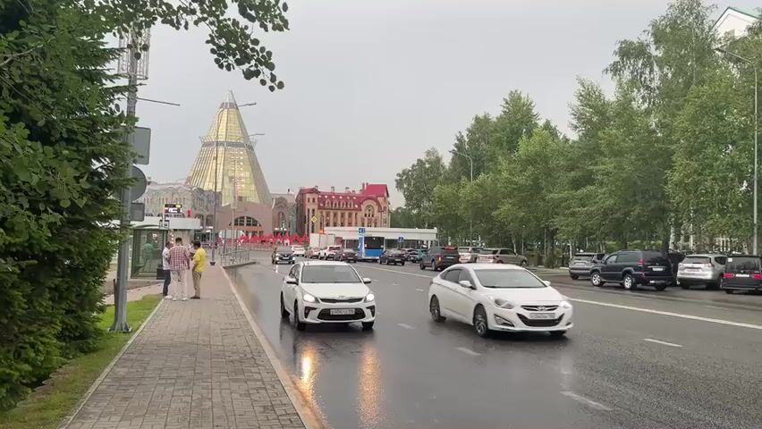 This media file depicts a street scene in Khanty-Mansiysk, Russia, under overcast skies. The road is wet, suggesting recent rain or ongoing drizzle. On the left side of the frame, a pedestrian walkway made of patterned tiles runs alongside a dense green hedge. A bus stop shelter is visible, with three men standing near it, appearing to be waiting or conversing.

The street itself is a multi-lane road with cars driving in both directions. In the foreground, a white Kia hatchback is moving forward, its tires reflecting the wet asphalt. Further down the road, a black Renault SUV is approaching. On the right side of the road, several cars are parked, including SUVs and sedans.

In the background, a distinctive building with a large, golden, conical roof dominates the skyline. Adjacent to it are other buildings with red roofs, suggesting a significant urban development. Tall green trees line the right side of the street, creating a natural border.

The overall mood is subdued, likely due to the cloudy weather. The scene suggests an ordinary day in an urban environment, with typical street activity. No specific event is evident, beyond the general movement of traffic and pedestrians. There is no visible text in the media file, except for the license plate on the white Kia, which is difficult to read clearly. The time of day appears to be daytime, given the ambient light.