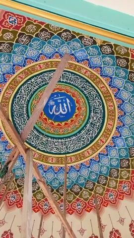 The video shows a low-angle view of an ornately decorated ceiling, likely inside a religious building such as a mosque. At the center of the ceiling is a large circular design featuring Arabic calligraphy in white against a backdrop of blue, red, and green concentric rings. Around this central circle is a more elaborate pattern of small, repeating designs with a variety of colors including blue, red, brown, and white. In the foreground, wooden scaffolding partially obstructs the view of the ceiling. The scaffolding is made of unfinished lumber and appears to be used for ongoing maintenance or construction work on the ceiling. The upper portion of the ceiling that is also visible above the circular design, is bordered by a light blue color.