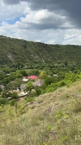 The video begins with a shot of a lush green valley with a village nestled among trees. The houses have red and white roofs. A steep, grassy hill slopes down to the right of the frame. The sky is partly cloudy. The camera then pans to the left, showing more of the valley and a person standing on the hill. The scene then shifts to a view from further up the hill, looking down at a path with people walking on it. A woman in a yellow dress and a child in an orange shirt are walking down the path, followed by another woman taking a photo. The path is lined with trees and grass, and there are houses visible in the distance. The camera continues to pan across the valley, showing more of the natural landscape and the village. The video ends with a wide shot of the valley, with the sun shining through the clouds.