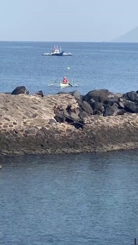 A video captures a coastal scene on a sunny day. In the foreground, a rocky breakwater dominates the view, its rough texture clearly visible. Beyond the breakwater, the calm blue sea stretches to the horizon. Two boats are present on the water. The closer boat is a smaller, traditional outrigger canoe with a person in a red shirt seated inside, seemingly paddling or engaged in activity. Further out, a larger, white boat with a red trim is anchored or drifting. In the distance, a hazy landmass or mountain range is visible against the clear sky. The water is a deep blue, and the sky is a pale, bright blue.