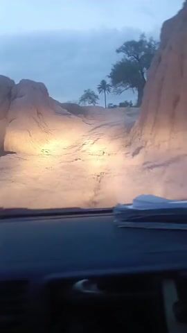 The video shows a car driving on a bumpy, unpaved road. The road appears to be sandy or made of light-colored dirt, with high banks on either side. Trees and some buildings are visible in the distance through the windshield. The sky is overcast. There is a stack of white papers on the dashboard of the car. The car is shaking due to the uneven road surface.  The headlights of the car are on, illuminating the road ahead.