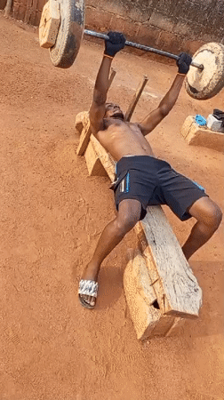 Man weightlifts on makeshift bench in outdoor Bukuru setting