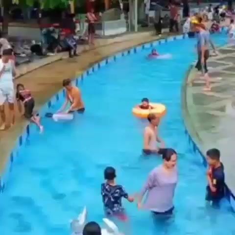 The video shows a crowded outdoor swimming pool with light blue water. Many people of various ages are swimming and playing in the water, while others are standing or sitting along the pool's edge. Some people are using inflatable tubes. Trees provide shade around the perimeter of the pool area. The atmosphere appears lively and recreational, with people enjoying their time at the pool.