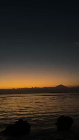The video captures a serene seascape at dusk. The sky transitions from a deep blue at the top to a warm orange glow near the horizon, indicating the last moments of daylight. A solitary star is visible in the dark blue expanse. In the distance, a silhouette of mountains, possibly a volcano, stands against the illuminated horizon. The sea below reflects the warm hues of the sky, with gentle waves lapping the shore. In the foreground, two dark, silhouetted rocks emerge from the water. The overall atmosphere is peaceful and tranquil, suggesting a quiet evening by the sea.
