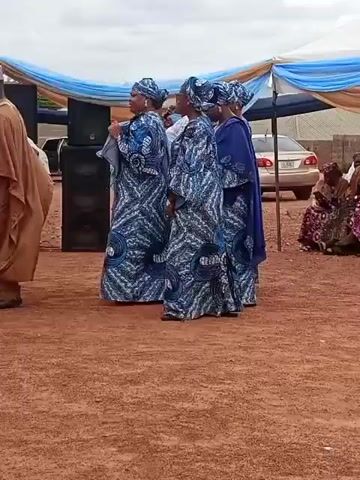 An outdoor social gathering takes place on reddish-brown dirt ground under an overcast sky. In the foreground, four women stand on the right, facing left, all wearing matching blue, white, and black patterned full-length dresses with coordinating headwraps. The woman on the far right also wears a solid royal blue shawl. Three men stand on the left, facing towards the women or the camera. One man wears a light-colored shirt and dark cap, another wears a dark brown traditional outfit (agbada) and cap, and the third wears a tan traditional outfit (agbada) and cap. A large speaker on a stand and a microphone stand are positioned between the two groups. In the background, two canopies draped with light blue and white fabric are visible, along with a white car and other people.