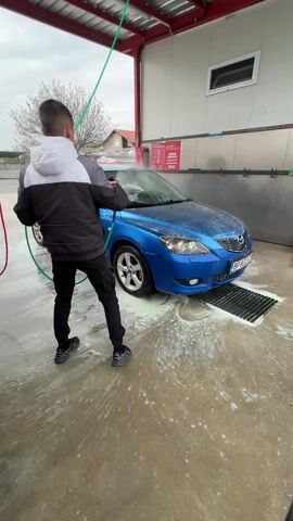 Here's a detailed description of the video:

The video shows a young man washing a blue Mazda 3 at a self-service car wash. He is standing behind the car, facing it, and holding a green high-pressure hose. He's wearing a dark jacket with a white hood, black pants, and dark sneakers. The car is covered in white foam. The floor around the car is wet and soapy.  A metal grate is visible in front of the car. 

The car wash bay has a red roof and grey wall with a window.  A sign with text is mounted on the wall. Red and green hoses hang from the ceiling. Beyond the car wash bay, trees and buildings are visible in the background. The sky is overcast.