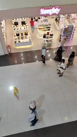 This is an aerial video shot from a high vantage point, looking down into a retail store and the surrounding mall area. The store, identified by the prominent sign "Naughty," appears to be a shoe and accessory shop, with displays of handbags and footwear visible. The store is brightly lit with white and pink fluorescent lights. A woman in a red shirt is seen inside the store, seemingly attending to customers.

Outside the store, in the mall walkway, several individuals are visible. On the left side of the frame, a person wearing a white shirt and dark pants is walking away from the camera, with another person in a white headscarf and blue jeans walking towards the camera. Further down, a person in a grey headscarf and a light-colored top is walking away from the camera, accompanied by a small child in a blue outfit.

In the center of the frame, a group of people are walking. One person, wearing a white t-shirt and shorts, is walking towards the camera, with a person in a black headscarf and abaya walking beside them. Behind them, another person in a black headscarf and a patterned dress is also walking.

A yellow sign is visible on the floor in the lower left quadrant of the frame. The floor is tiled with large, light-colored square tiles. The lighting in the mall area is dimmer than inside the store, with reflections visible on the floor. The overall scene depicts a typical day in a shopping mall.