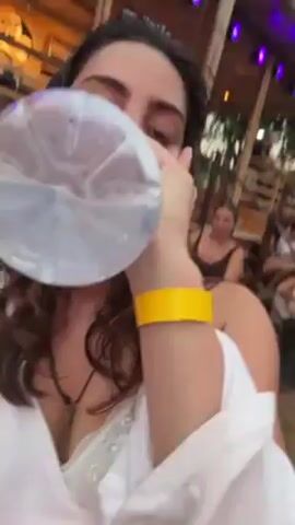 A person wearing a white shirt and a yellow wristband is holding a clear plastic water bottle with a blue label. The bottle appears to have condensation on it. The camera angle is slightly angled. The background shows a sandy ground, blurred figures of people, and a canopy with colorful decorations. The person holding the bottle appears to be at an outdoor event.