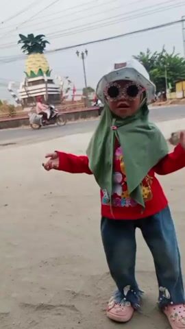 A young child wearing a red long-sleeved shirt, blue jeans, a green hijab, a white bucket hat, and large, sparkly sunglasses is dancing in front of a dirt road. The child's jeans have distressed fringe at the bottom, and they are wearing pink sandals. In the background, there is a large pineapple monument, a street with some vehicles, and trees. The sky is overcast. The child appears to be looking at the camera and moving their arms.