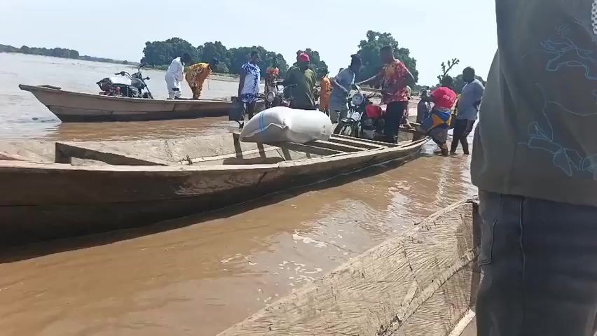 Several people are loading items onto a small, wooden boat in shallow, muddy water. One boat already has a white bag loaded on it, while another is being loaded with a motorcycle.  There is another motorcycle already loaded onto the first boat. Some people stand in the water near the boats, appearing to assist with the loading. The camera is positioned on another boat. A young boy is shown pushing the boat and a man in jeans appears to be steering the boat into deeper water. The water is brown and murky. The background shows a landscape with trees. The sky is bright and clear.