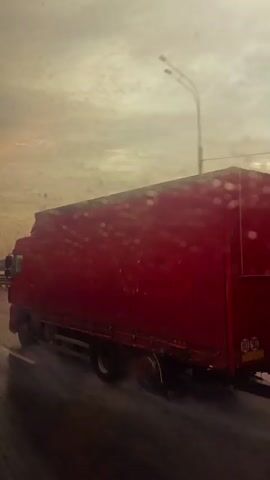 The video shows a red semi-truck driving on a wet road with a hazy, overcast sky. Raindrops are visible on the camera lens, obscuring the view slightly. Several other vehicles, including a blue car, are also present on the road, driving in the same direction. The truck is in the foreground, taking up a significant portion of the frame. The road appears to be an elevated highway, with a guardrail visible on the left side. The overall atmosphere is somewhat grim and muted due to the weather conditions.