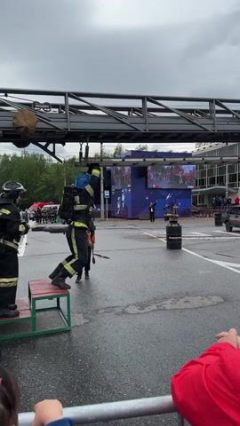 This video captures a dynamic scene outdoors. In the foreground, spectators, identified by their red clothing and hands, are visible behind a railing, implying an event or demonstration is taking place. The central action involves two individuals dressed in firefighter gear. One firefighter stands on a green and red platform, looking towards the other. The second firefighter is mid-air, suspended by a wire from a large metal structure resembling a bridge or an elevated walkway. This firefighter is actively engaged in a maneuver, appearing to swing or jump. A third person, dressed in athletic wear with a jacket, stands between them, potentially acting as an instructor or demonstrator, holding a tool. In the background, a large screen displays images, and other people and vehicles are present, suggesting a public gathering or competition. The ground is wet, indicating recent rain or a damp environment. The sky is overcast.