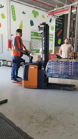 The video shows three men in a warehouse setting. One man, wearing a blue shirt and an orange safety vest, is operating a yellow and black forklift. He is standing on the forklift's platform and holding the controls. To his right, a man in a tan t-shirt is standing and appears to be talking. Further to the right, another man in a light-colored t-shirt is standing and looking towards the forklift operator. The forklift is carrying a pallet of boxed items wrapped in plastic. The warehouse has a concrete floor and metal shelving in the background. There are colorful graphics on the wall to the left, depicting food items and icons. The lighting is bright, suggesting an indoor environment. The overall scene depicts a routine operation within a warehouse or distribution center.