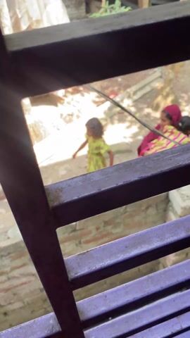 The video is shot from a slightly elevated perspective, looking down into an outdoor area. The foreground is dominated by a dark, possibly purple-hued, metal railing or grate, partially obscuring the view. Bright sunlight streams into the scene, casting strong shadows.

In the background, there is an unpaved, dusty ground with visible soil and scattered debris like small rocks and what appear to be pieces of plastic. Several tree trunks, resembling palm trees with visible root systems, are scattered across the area. A white tarp or plastic sheeting is seen bunched up near the base of one of the trees. A low brick wall, constructed from reddish-brown bricks with mortar, runs horizontally across the mid-ground.

At the beginning of the video (0:00), two figures are visible behind the metal railing and above the brick wall. One is a small child, wearing a light green and yellow patterned outfit, standing. The other is an adult, likely a woman, wearing a dark red garment (possibly a head covering or part of a traditional dress) and a brightly colored top with pink and yellow stripes or patterns. The adult is initially seen in a crouched or seated position.

Around 0:01, the adult rises and is seen holding or embracing the child, who is now clearly wearing a pink, green, and yellow striped top. The adult's red garment covers her head and shoulders. They are standing behind the low brick wall. The camera slightly adjusts, maintaining focus on them.

The adult and child then begin to walk together towards the right side of the frame (0:02). The child appears to be leaning against or being carried by the adult, with their face mostly obscured. The adult's face becomes slightly more visible as they move, though not clearly defined.

By 0:04, the adult and child move out of the frame to the right. The video then focuses on the ground, the brick wall, and the roots of the trees until it ends.

Throughout the video, faint human voices are audible, suggesting conversation or interaction happening nearby, but the specific words are not discernible. The overall impression is that of a candid, everyday moment captured from an elevated viewpoint.