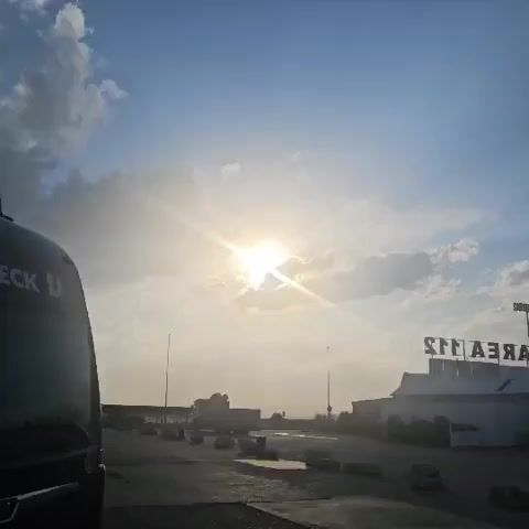 A wide shot captures a sunlit scene, with the sun prominently featured in the upper center of the frame, partially obscured by clouds. The sky is a bright blue, transitioning to a hazy yellow near the horizon. The sun's rays create a lens flare effect, radiating outwards. In the foreground, the left side of the image shows the front of a dark-colored vehicle, possibly a bus, with the partial text "ECK U" visible on its side. The ground in front of the vehicle appears to be a paved area, with scattered concrete barriers or large stones. In the background, a building with a sign that reads "ARENAS" in reverse is visible on the right. Several poles with lights are also present in the scene, spaced across the landscape. The overall impression is of an outdoor location, possibly a parking area or rest stop, bathed in the bright light of the sun.