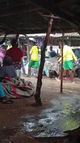 A group of people are gathered under a makeshift shelter with a corrugated metal roof. It appears to be raining, as water is visible on the ground and falling from the roof. Some people are wearing bright yellow shirts and green shorts, suggesting they might be part of a team or group. Others are wearing more casual clothing. In the foreground, a large, patterned bag is visible. The ground is muddy and wet, with puddles of water. The scene suggests a community gathering or market taking place outdoors despite the weather.