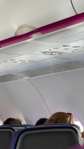 The video shows the interior of an airplane cabin from a passenger's perspective. The camera is pointed upwards towards the overhead compartments and lighting. Several passengers are visible from the back of their heads, seated in rows of airline seats. The overhead compartment bins are open and partially obscured by the curvature of the cabin ceiling. There are overhead lights and signs visible, including a "no smoking" sign with a lit cigarette symbol crossed out. A pink trim runs along the edge of the overhead compartments. A faint mist or vapor appears to be emanating from the overhead vents, suggesting the air conditioning or cabin pressurization system is active. The overall environment appears to be typical of a commercial airplane during flight or boarding.
