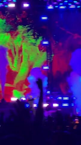 A concert is in progress. A male singer is on stage, holding a microphone, and singing. He is wearing a dark outfit. Blue and purple smoke effects are being used on stage, obscuring parts of the singer. The stage backdrop is a large screen displaying abstract, colorful graphics with red and yellow splashes. Stage lights are visible above the screen. A drummer and guitarist are also on stage, partially visible in the background. Silhouettes of an audience are visible in the foreground, with hands raised in the air.