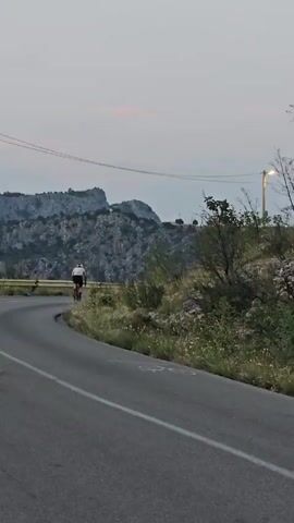 The video shows a cyclist riding on a winding road through a mountainous, rocky landscape. The cyclist is wearing a helmet and a white jersey with red shorts. The road is paved and has white markings on it. There are trees and bushes on the side of the road, and the mountains in the background are rocky and sparsely vegetated. The sky is overcast.