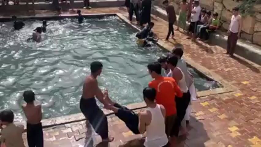 The video shows a large group of males of various ages swimming and playing in a rectangular pool outdoors. Some individuals are throwing others into the water. Several people stand around the perimeter of the pool, some clothed in traditional attire, observing the activity. The pool is surrounded by brick paving and stone walls.  Some green foliage is visible in the background. The scene is lively and appears to be a recreational event.