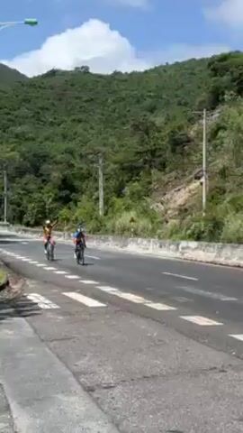 The video shows two cyclists riding road bikes on a paved road alongside a mountainside. The cyclists are wearing matching red, yellow, and blue cycling kits and helmets. They are riding in the same direction and are close to each other. A white van passes them in the opposite direction. The camera moves to show a white car parked with its driver-side door open. The background consists of a lush green mountainside covered with trees and vegetation. The sky is overcast. Traffic signs are visible along the road.