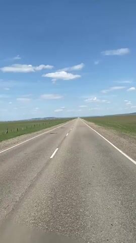 The video shows a straight, two-lane road stretching to the horizon under a bright blue sky with scattered white clouds. Green fields line both sides of the road, separated by a fence. In the distance, a town or settlement is visible. The camera appears to be mounted on a vehicle traveling down the road. There is music playing.