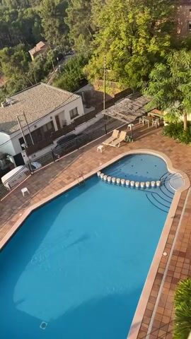 The video shows a swimming pool with clear blue water. The pool has a kidney shape and is surrounded by a tiled patio area. There are several white lounge chairs on the patio, as well as some white chairs and tables under a shaded area. The pool area is also bordered by a fence and some greenery, including trees and bushes. In the background, there are some houses and a hillside covered in trees. The sun is shining, casting shadows on the water and patio. There is no visible activity in or around the pool at the beginning of the clip, but at 00:02 a person can be seen entering the water from the steps on the right side of the pool. They appear to be wearing a dark swimsuit and move into the pool.
