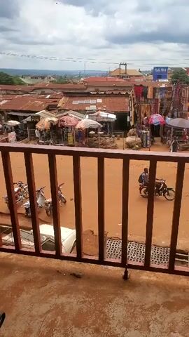 The video shows a view of a bustling marketplace from behind a brown metal railing. The ground visible through the railing is a reddish-brown color. Beyond the railing, there is a dirt road with people walking and riding motorbikes. The marketplace is filled with stalls and vendors selling various goods under umbrellas and canopies. Buildings with red and white roofs are visible in the background. The sky is overcast with grey clouds.