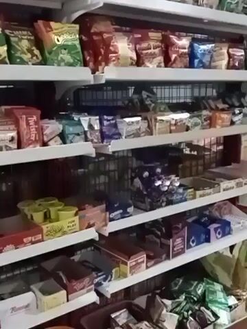 The video shows shelves stocked with various snacks and food items. The shelves are white and metal, and they appear to be in a store or a pantry. The items on the shelves include bags of chips, candy bars, cookies, and other packaged goods. Some of the brands visible include Cheetos, Twix, and Oreo. The lighting in the scene is somewhat dim, but the items are clearly visible. The camera is positioned to provide a full view of the shelves.