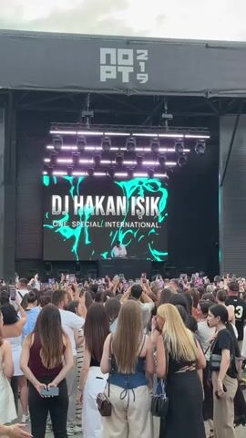 A large crowd of people are gathered in front of an outdoor stage. A large screen behind the stage displays the text "DJ HAKAN ISIK" and "ONE. SPECIAL. INTERNATIONAL." A DJ is visible on stage behind a console, with lights illuminating the area. The crowd is holding up their phones, presumably to record or take pictures of the performance. The overall atmosphere appears to be one of excitement and enjoyment.