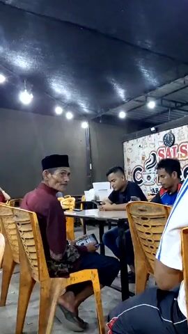 The video shows four men sitting at tables in an indoor café or restaurant. The man closest to the camera wears a maroon shirt, black pants, a black peci hat, and slippers. He is looking to his right and talking, while holding a tablet. Another man sits across the table from him, looking at a smartphone. To his right, a third man in a blue shirt looks on. The fourth man is partially visible on the right, wearing a white shirt and black pants. The café is dimly lit by several visible overhead lights. The tables are black and the chairs are yellow. A wall in the background has a mural with coffee-related images and the words "Salsa Coffee".