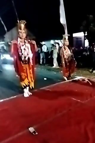 Two individuals, dressed in elaborate traditional costumes and headwear, stand facing the camera on a red carpet. They are outdoors at night, with an audience visible in the background. The individuals are adorned with colorful fabrics, gold ornamentation, and what appear to be sashes or scarves draped around their necks and arms. The person on the left is wearing a red outer robe over a patterned tunic and skirt, while the person on the right wears a similar outfit with predominantly yellow and red tones. Both individuals have their hands positioned in a way that suggests they are performing a dance or ritual. The environment is dark, with some lighting from unseen sources illuminating the scene. Flags are visible in the background, and a white line is marked on the ground in front of the carpet.