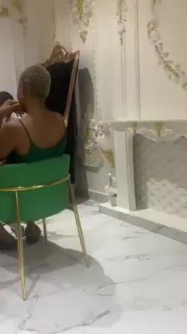 Several individuals are seated in a room with ornate white walls and a marble floor. Two of them are in the foreground sitting on green chairs with gold frames. One wears a green tank top and the other a red and white shirt. One individual takes their shoes off.
