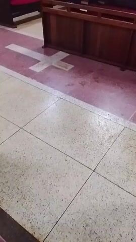 The video shows the floor of what appears to be a church interior. The floor is primarily composed of light-colored, speckled tiles, with dark lines marking the tile edges. In the background, there is a section of the floor in a reddish-purple hue, featuring a large white cross design. Next to this section is a dark wooden church pew. The camera angle is stationary, looking down at the floor. The lighting is moderate and indoor. There are no visible people in the shot.