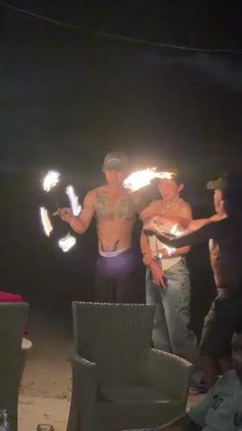 A shirtless man with tattoos and a baseball cap spins two flaming poi in front of him. To his right, a woman wearing a light-colored strapless top and light pants holds a burning object, possibly a small fire bowl or stick, while smiling and looking towards the man. Another person, partially visible and lower to the ground, reaches towards the burning object held by the woman. The scene is dark, indicating nighttime, with the fire being the primary light source, and several chairs are visible in the foreground.