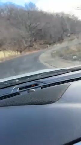 The camera is inside a vehicle, recording the view through the windshield. The vehicle is driving on a winding, paved road. Trees line both sides of the road, and their branches are mostly bare. In the distance, a mountain or hill is visible under a sky with scattered clouds. The dashboard of the car is visible in the foreground, including a speaker. The sky becomes brighter as the vehicle moves.