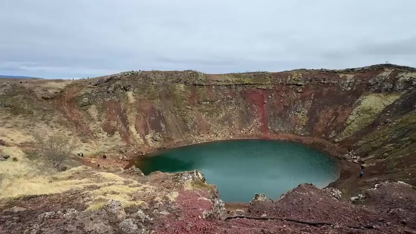 The video shows a wide shot of a volcanic crater filled with turquoise-colored water. The crater walls are reddish-brown and have some green vegetation. The sky is overcast. There are a few people visible at various points on the crater rim and along a path descending into the crater. The foreground shows rocky terrain.