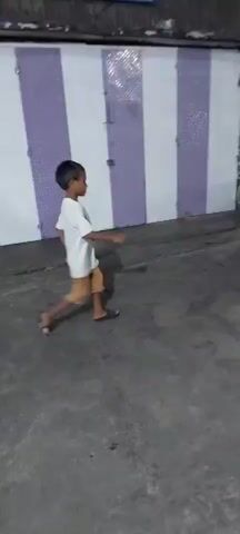 Two young boys are playing with a soccer ball in what appears to be an alley or driveway at night. The boy on the left is wearing a white t-shirt and tan shorts. The boy on the right is wearing a dark blue shirt with graphics and dark shorts. They are kicking the ball back and forth. In the background, there is a building with purple and white striped doors or shutters. There are also motorcycles parked on the right side of the frame. The ground is paved and appears to be wet. The lighting suggests it is nighttime, with artificial lights illuminating the scene.