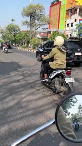 The video shows a street scene with several motorcycles and cars. The camera is positioned behind a motorcycle, with its side mirror visible in the foreground. In the middle ground, a person wearing a helmet and a tan jacket is riding a white scooter. The scooter has the license plate W 6528 AC. To the right of the scooter, a black car is visible. The background shows buildings and trees, with a clear blue sky. The road is paved and appears to be in good condition. The lighting suggests it is daytime, with shadows cast on the road.