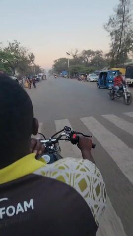 The video, taken from the perspective of a rider on a motorcycle, shows a street scene in what appears to be a tropical or semi-tropical locale. The rider, wearing a black and white patterned shirt with a yellow collar, grips the handlebars as they navigate the road. The street has a fair amount of traffic, including other motorcycles, cars, a blue auto-rickshaw (tuk-tuk), and a truck in the distance. Pedestrians are visible on the side of the road and in areas where motorcycles are parked. Trees line the sides of the street, and a solar-powered streetlight is visible. The sky is light, indicating that it is either early morning or late afternoon. The overall impression is of a busy street in a developing country.