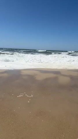 The media file depicts a scenic view of the Atlantic Ocean from a sandy beach in Torreira, Portugal. The scene is set on a bright, sunny day, with a clear, vivid blue sky dominating the upper portion of the frame. The ocean is active, with moderate waves rolling towards the shore, creating a white foamy surf that gently washes onto the wet sand. The wet sand reflects the sky and the waves, creating interesting patterns of light and shadow. Small bubbles or foam formations are visible on the sand in the foreground. There are no people or objects clearly visible in the scene. The overall mood is one of tranquility and natural beauty, characteristic of a peaceful beach day. The time of day appears to be midday, given the brightness of the sun and the clear sky. The location is confirmed as Torreira, Portugal, known for its beautiful beaches along the Atlantic coast. There is no visible text in the media file.