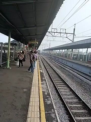 The video captures a view of a train station platform on a bright day. On the left side of the frame, a covered platform area extends into the distance. Several individuals are visible on the platform, including a woman holding a child, and another woman carrying a bag. Some people are standing, while others are sitting. Further down the platform, a man in a grey shirt and blue jeans stands near the edge, looking towards the train tracks. The train tracks themselves are prominent in the right side of the video, running parallel to the platform. Overhead power lines are visible above the tracks. A train is faintly visible in the distance on the tracks. The platform appears to be made of concrete, with yellow tactile paving along the edge. Fencing is visible behind the people on the platform, and what looks like parked motorcycles can be seen behind the fence. The sky is overcast, suggesting a cloudy or hazy day.