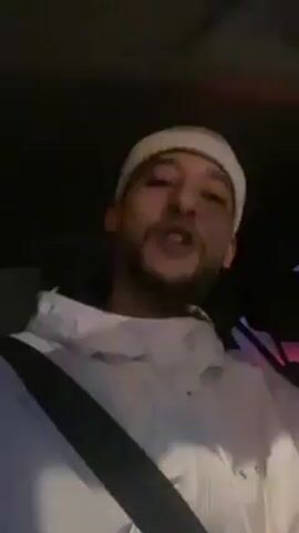 A man is recording a video of himself inside a car at night. He is wearing a light-colored beanie and a white hoodie. The car’s interior is dark, and he is wearing a seatbelt. He is gesturing with his hands and singing or rapping along to music playing in the car.